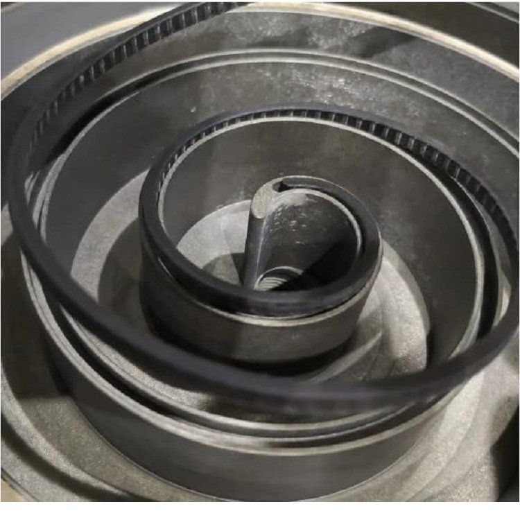 The image depicts a close-up view of an industrial mechanical assembly featuring a spiral-shaped, toothed timing belt coiled around a series of gray metallic pulleys. The belt, likely made of rubber or a similar material, has a ribbed or serrated surface designed for enhanced grip and precision. The pulleys are stacked in a layered configuration, with a central cylindrical component possibly serving as the main shaft or drive mechanism. The overall setup suggests a component used in machinery, such as a conveyor system or engine, with visible wear and dust indicating prolonged use.